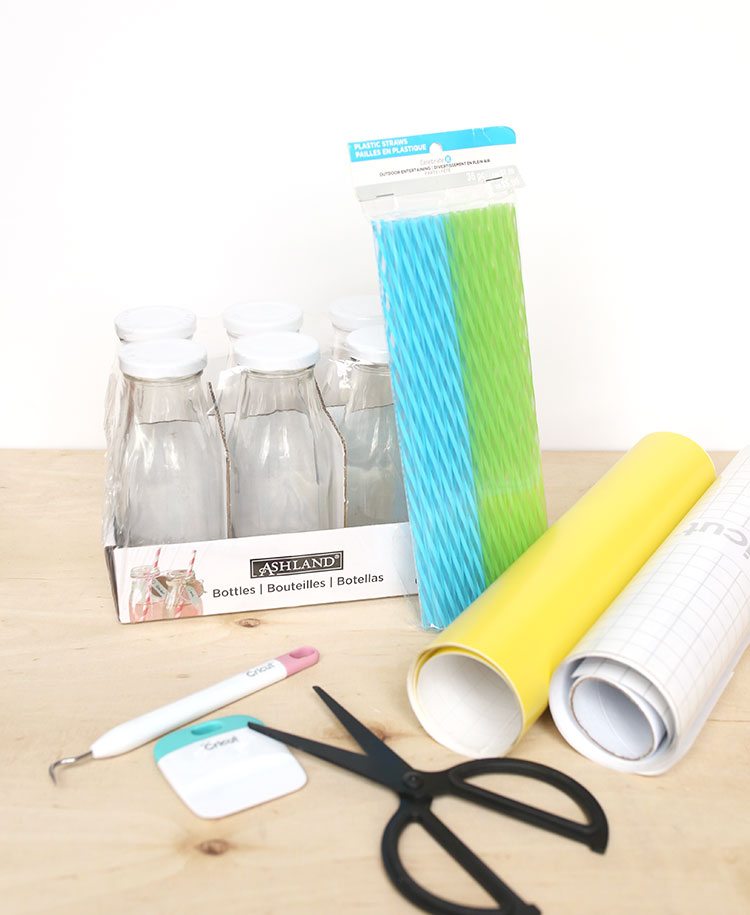 Craft supplies on a wooden background: scissors, plastic straws, vinyl, Cricut tools, and glass milk bottles