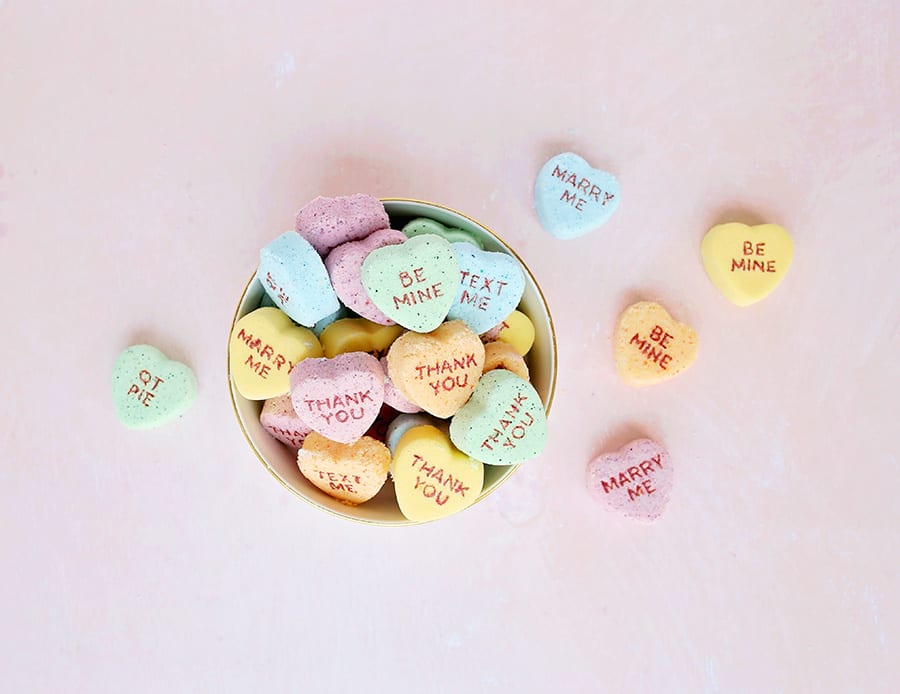 Yellow, orange, blue, and green conversation heart Valentine's Day bath bombs in a bowl on a pink background