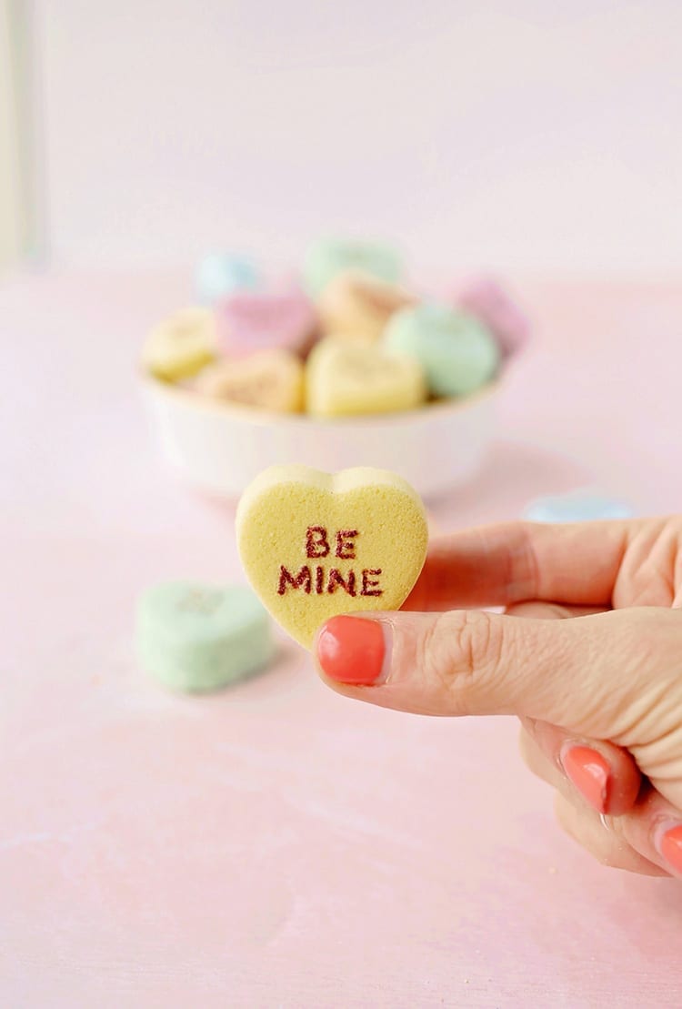 Yellow, orange, blue, and green conversation heart Valentine's Day bath bombs in a hand on a pink background