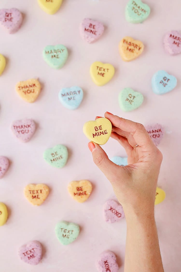 Yellow, orange, blue, and green conversation heart Valentine's Day bath bombs in a hand on a pink background