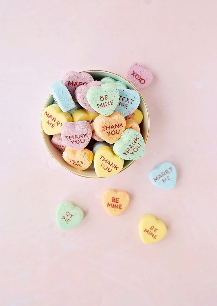 Yellow, orange, blue, and green conversation heart Valentine's Day bath bombs in a bowl on a pink background