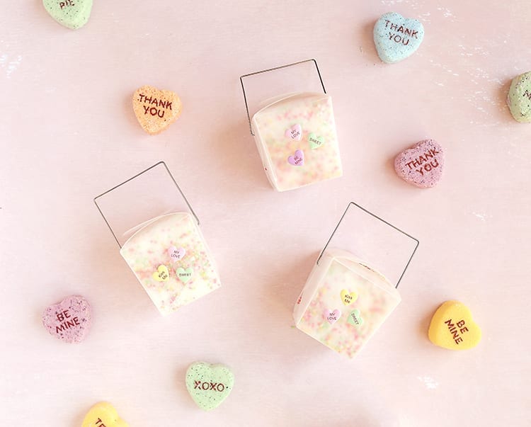 Clear takeout containers of valentines day bath salts on a pink background with giant conversation hearts