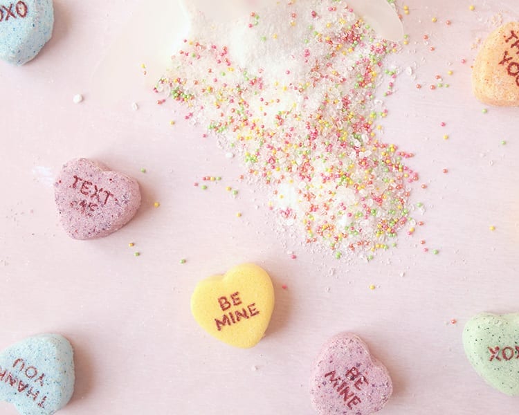 Conversation hearts Valentine's Day bath salts with colorful sprinkles spilled onto a pink background with giant conversation hearts