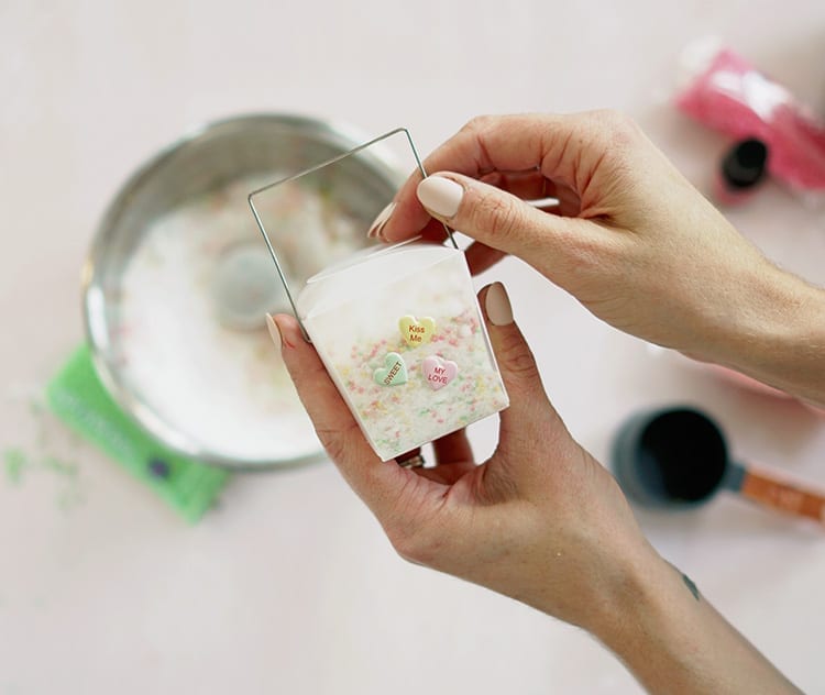 Caucasian hands holding a clear takeout container with conversation hearts stickers and containing a Valentine's Day bath salts recipe mixture