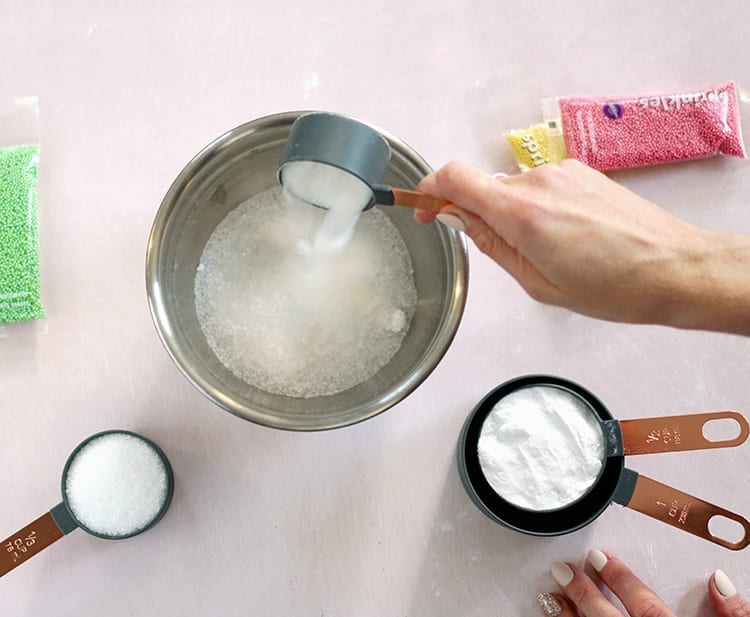 Caucasian hand adding a grey measuring cup of salt to a metal mixing bowl on a pink background