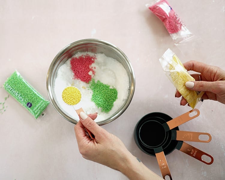 Caucasian hand pouring a spoonful of yellow sprinkles into a metal mixing bowl on a pink background