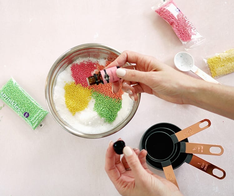 Hand adding a drop of cotton candy scented oil to Valentines Day bath salts recipe mixture in a metal bowl on a pink background