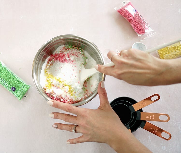 Caucasian hand mixing Valentine's Day bath salts recipe in a metal bowl with a white spoon over a pink surface