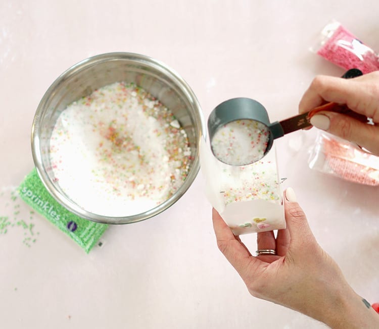 Caucasian hand pouring Valentine's Day bath salts recipe mixture from a grey measuring cup into a clear takeout box over a pink background