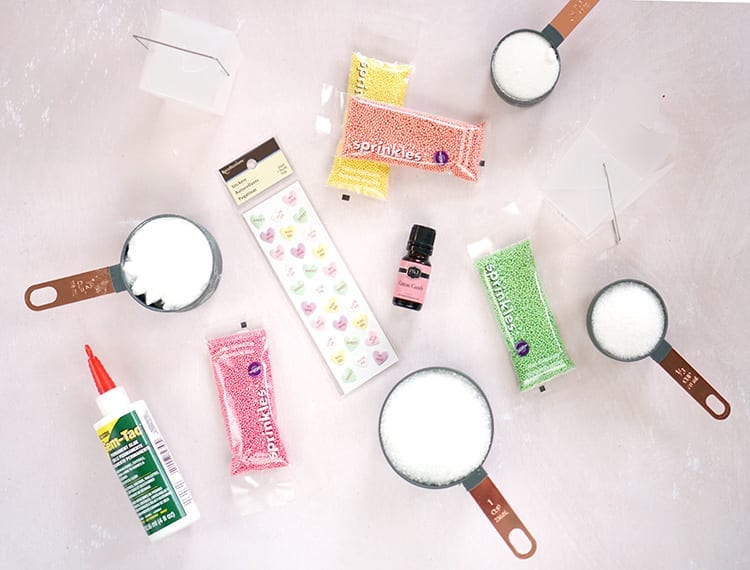 Colorful sprinkles, conversation hearts stickers, measuring cups full of white powder ingredients, and glue on a pink background