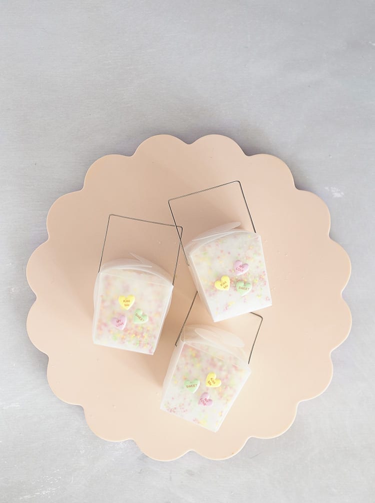 Three clear takeout containers with conversation hearts stickers on a pink scalloped tray on a grey background