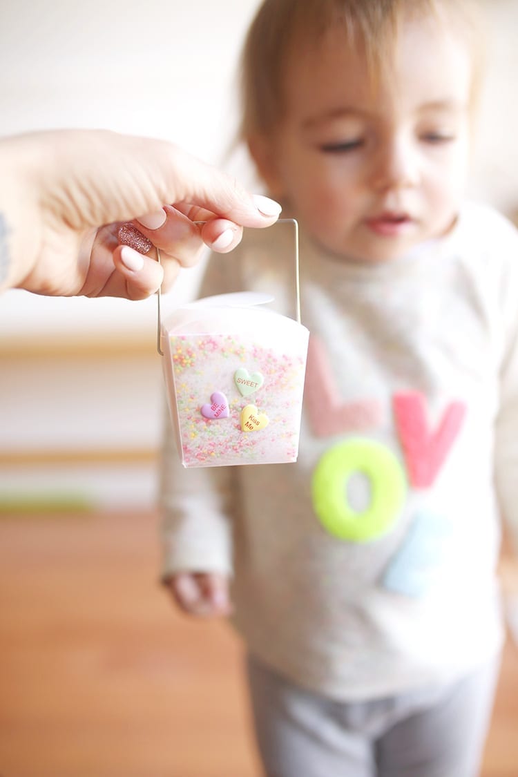 Hand holding a clear takeout container of Valentine's Day bath salt recipe in front of a baby wearing a sweatshirt that says LOVE