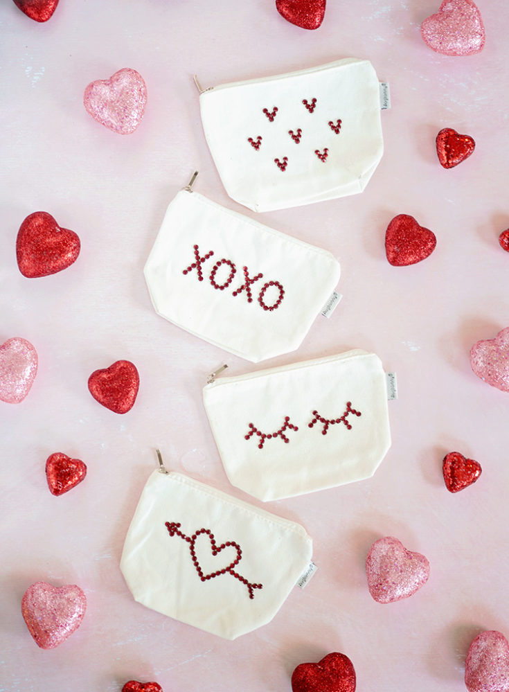 Four white canvas pouches with red rhinestone assorted designs. Lying on a pink background with pink and red glitter hearts scattered around