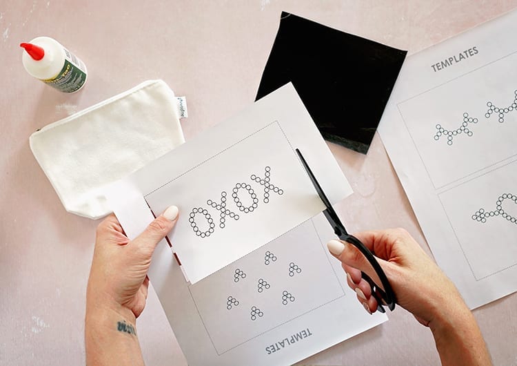 Caucasian hands cutting out an XOXO template from white paper with black scissors against a pink background