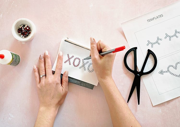 Caucasian hands tracing XOXO template with red pen on white paper against a pink background