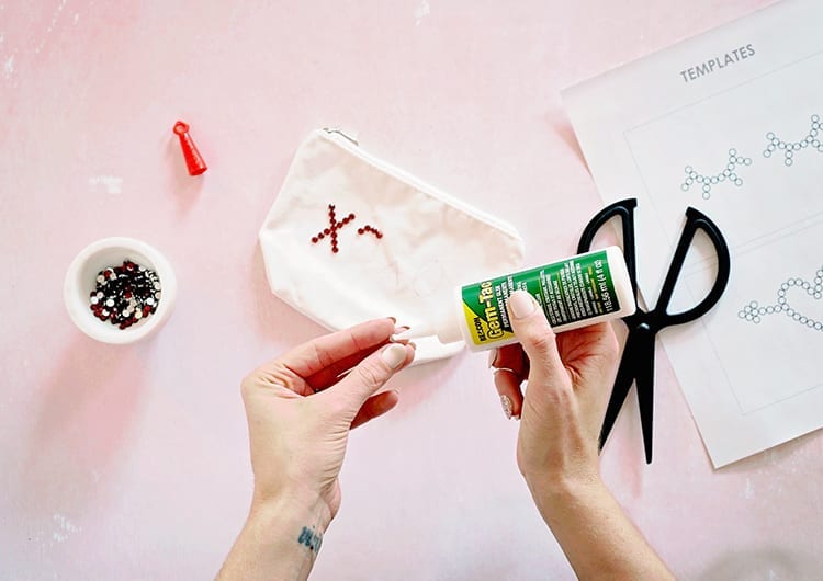 Hands applying Beacon Adhesives Gem-Tac to a red rhinestone against a pink background