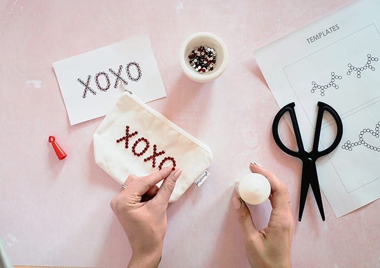 Hands applying a red rhinestone to an XOXO design on a white canvas pouch against a pink background