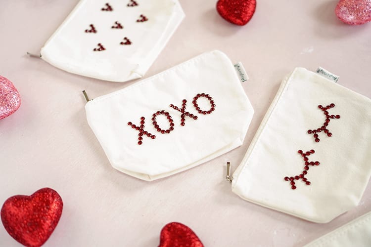 Four white canvas pouches with red rhinestone assorted designs. Lying on a pink background with pink and red glitter hearts scattered around