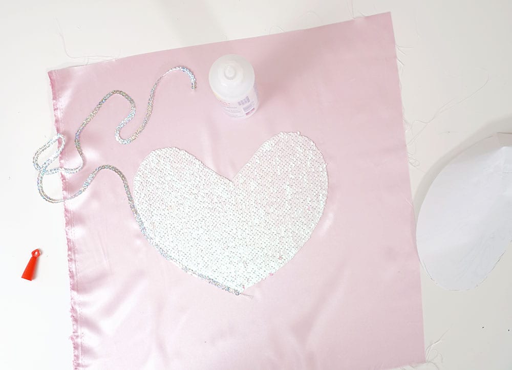 Bottle of Beacon Adhesives Fabri-Tac glue on top of a mermaid sequin fabric heart and pink satin to make a magic sequin pillow