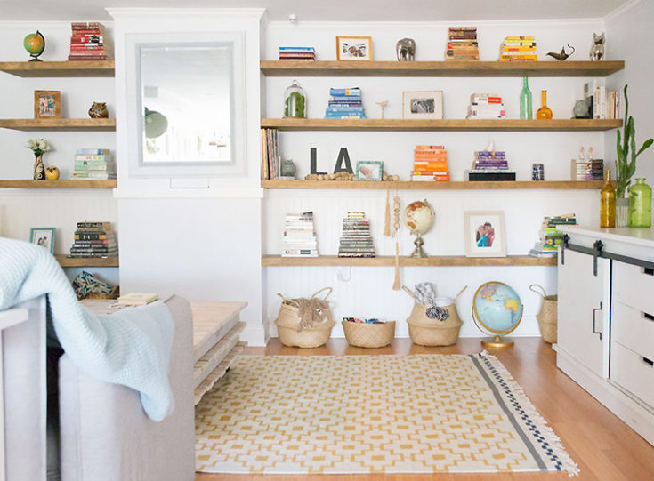 Wall of wooden floating shelves with books and knick knacks on them. Baskets and a globe on the floor in front of a yellow and white patterned rug.