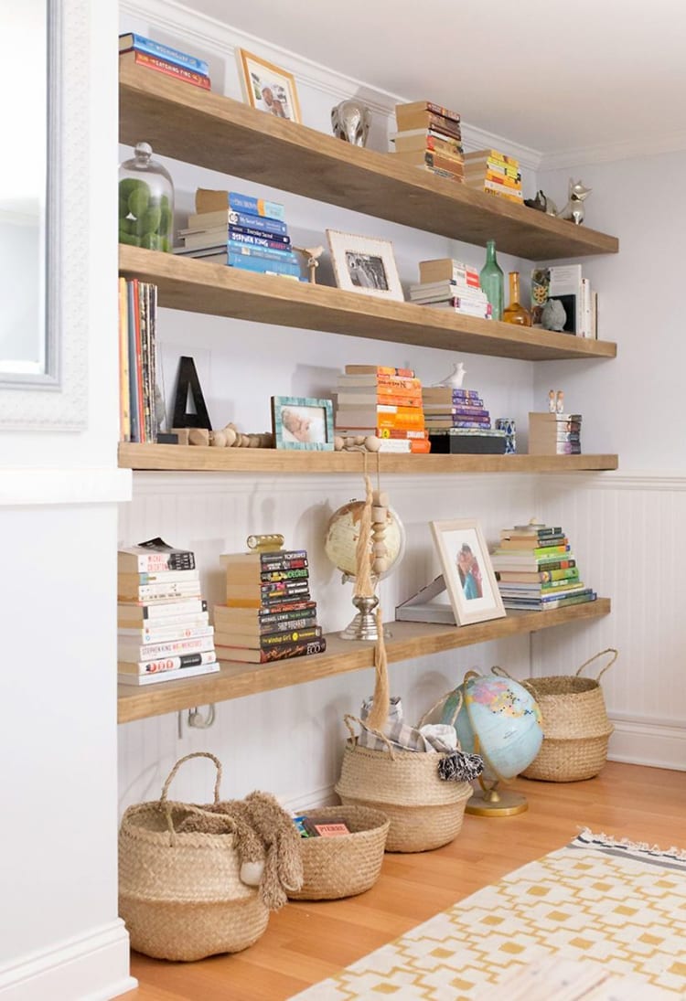 Wall of wooden floating shelves with books and knick knacks on them. Baskets and a globe on the floor in front of a yellow and white patterned rug.