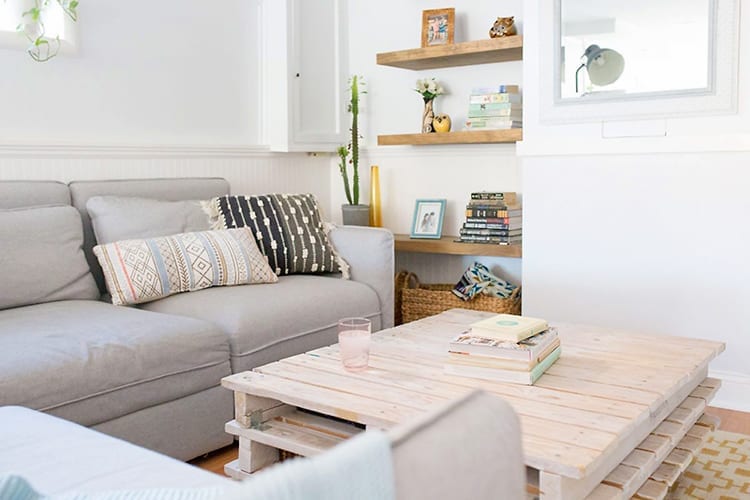 White living room with light grey sectional sofa, whitewashed wooden pallet coffee table, and wooden floating shelves