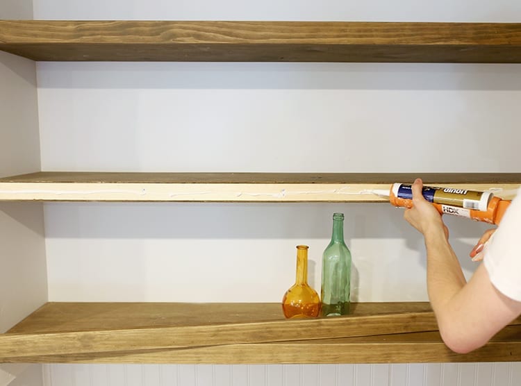 Man gluing the front of a floating shelf against a very light blue wall