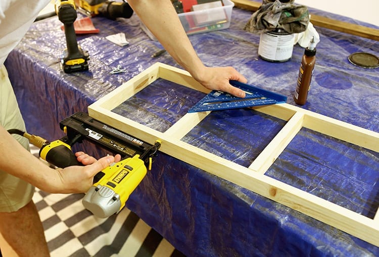 Caucasian hands drilling a screw into a wooden shelf frame on a table covered in a blue tarp