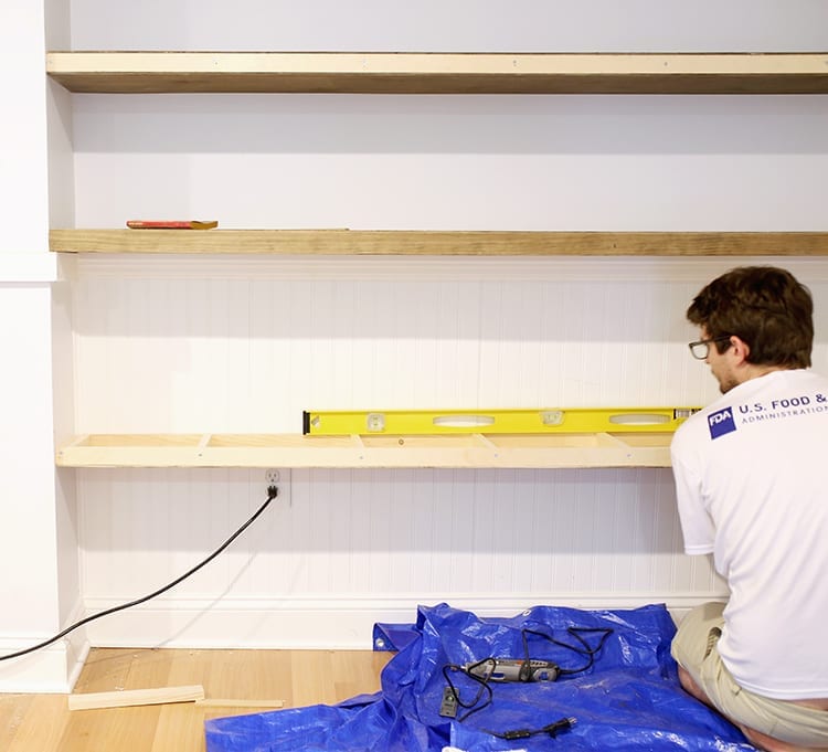 Man installing a pine shelf frame onto a white wall over a blue tarp