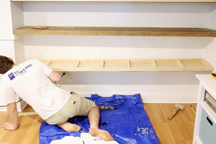 Man installing a pine shelf frame onto a white wall over a blue tarp