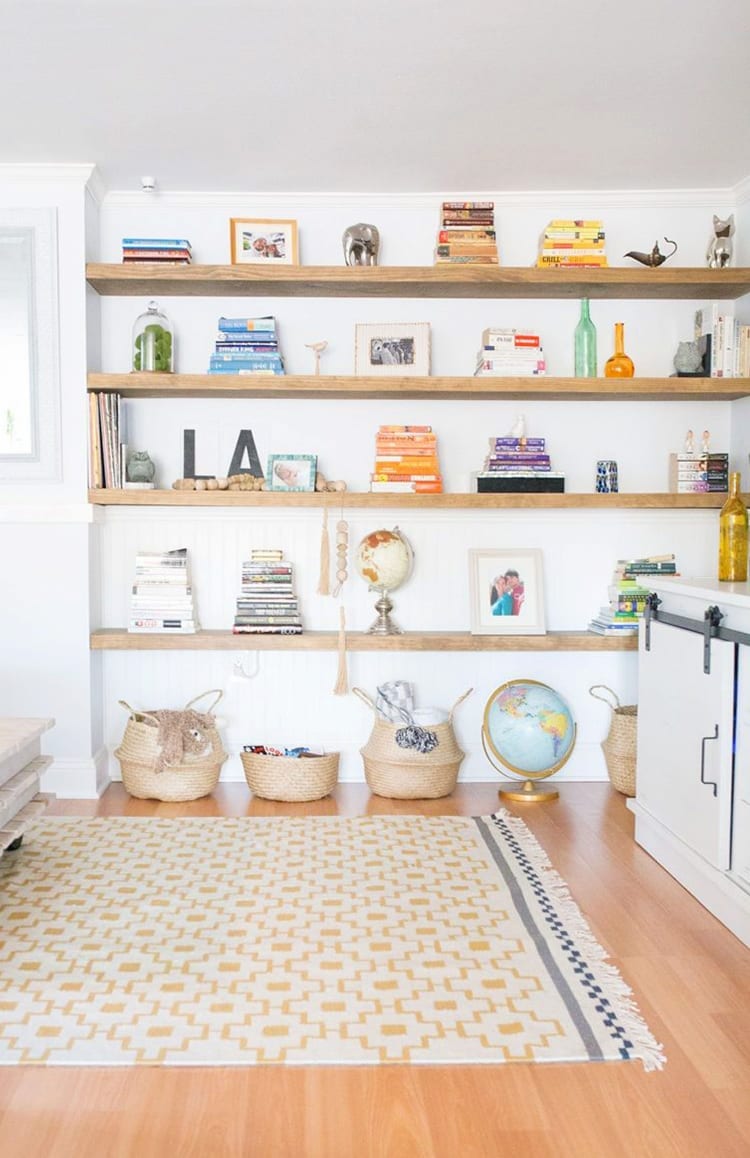 Wall of wooden floating shelves with books and knick knacks on them. Baskets and a globe on the floor in front of a yellow and white patterned rug.