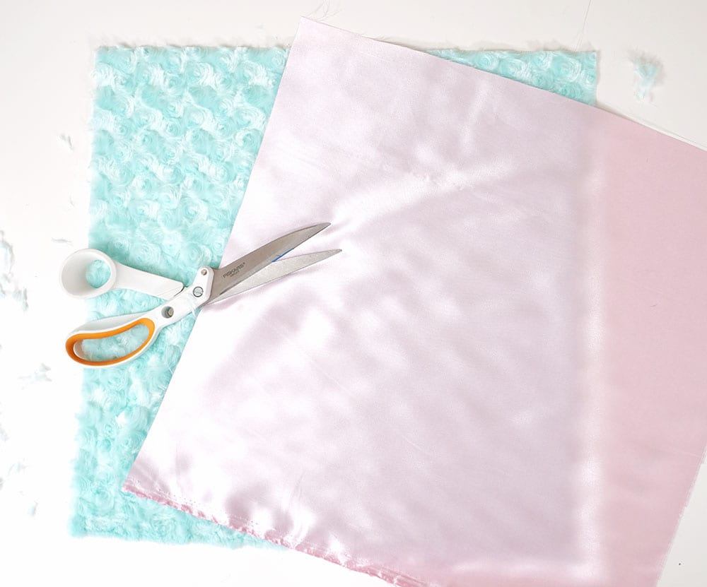 Sewing scissors on top of magic sequin pillow supplies: pink satin and turquoise faux fur fabric squares on a white background