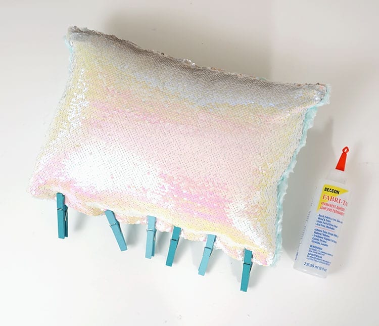 Mermaid sequin pillow with blue clothespins along the bottom and a bottle of Beacon Adhesives Fabri-Tac glue to the right