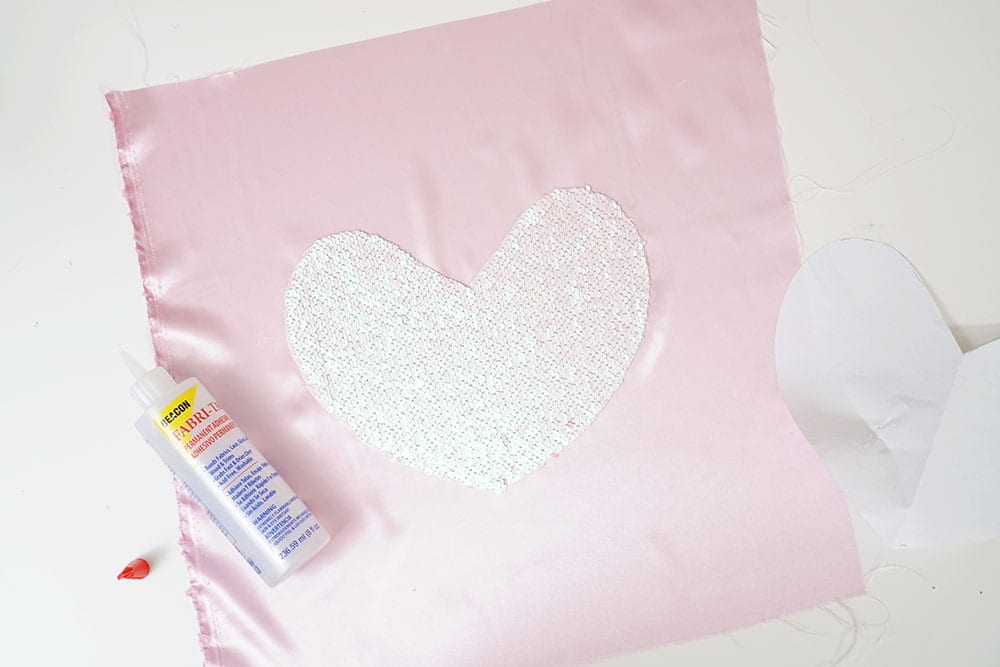 Bottle of Beacon Adhesives Fabri-Tac glue on top of a mermaid sequin fabric heart and pink satin to make a magic sequin pillow