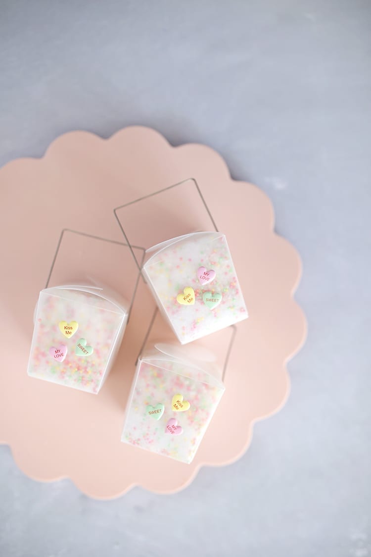Three clear takeout containers with conversation hearts stickers on a pink scalloped tray on a grey background