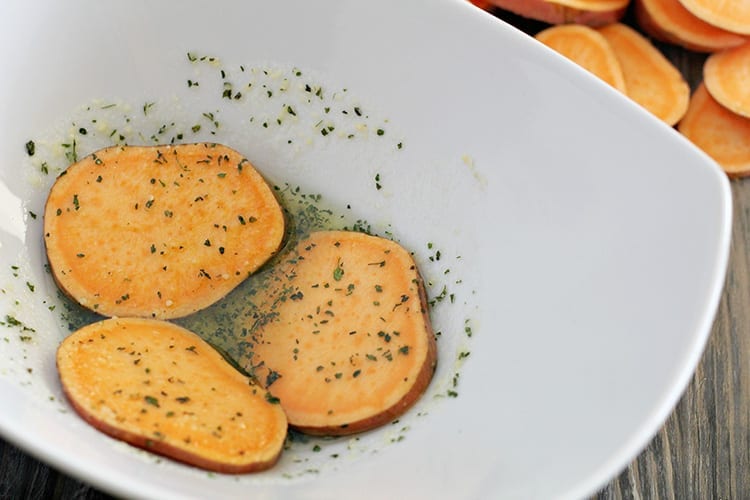How to Make Crispy Baked Sweet Potato Chips with Parmesan and Herbs with fresh organic sweet potatoes marinating in a white bowl with herbs