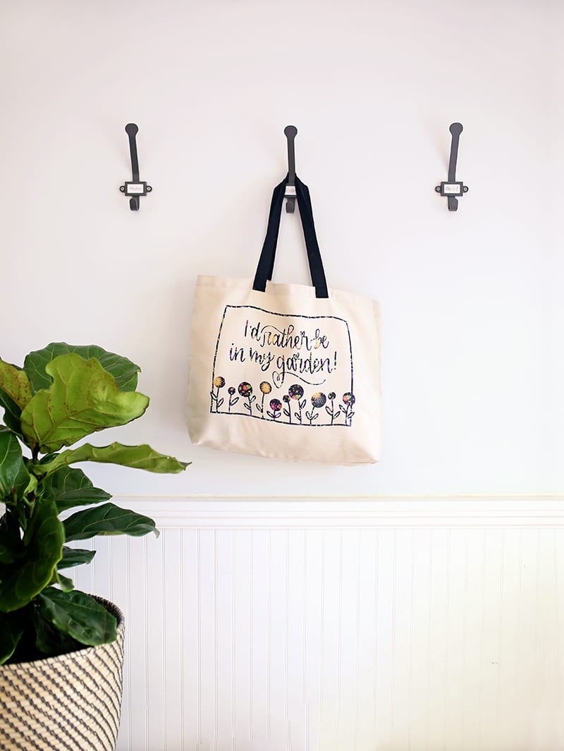 Gardener tote bag made with Cricut Infusible Ink Transfer Sheets hanging on a wall hook with a straw hat and fiddle leaf fig tree