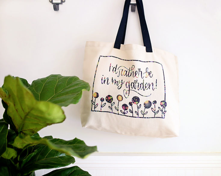 Gardener tote bag made with Cricut Infusible Ink Transfer Sheets hanging on a wall hook with a straw hat and fiddle leaf fig tree