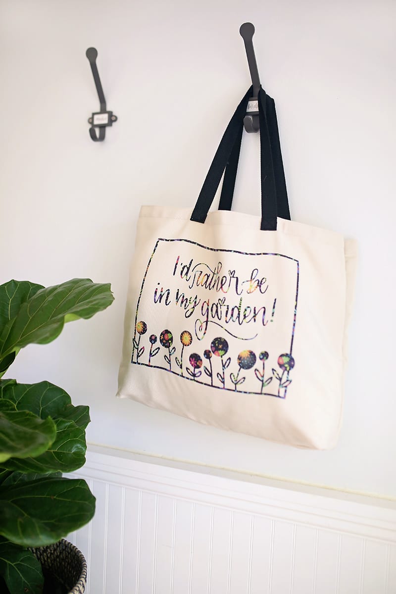 Gardener tote bag made with Cricut Infusible Ink Transfer Sheets hanging on a wall hook with a straw hat and fiddle leaf fig tree