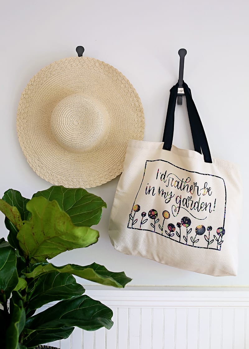 Gardener tote bag made with Cricut Infusible Ink Transfer Sheets hanging on a wall hook with a straw hat and fiddle leaf fig tree