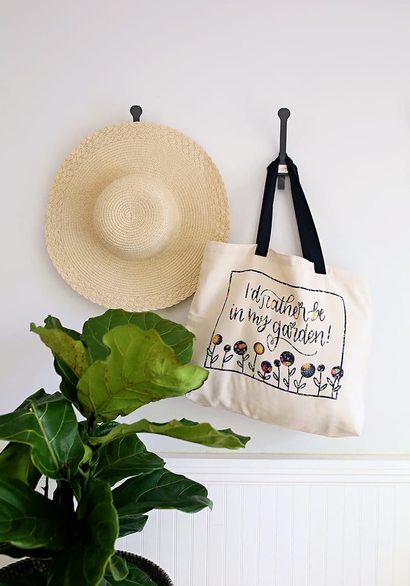Gardener tote bag made with Cricut Infusible Ink Transfer Sheets hanging on a wall hook with a straw hat and fiddle leaf fig tree