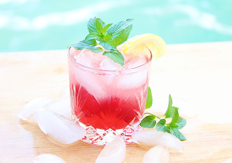 Cocktail in a rocks glass with cherry lemonade drink and a sprig of mint next to a pool for summer