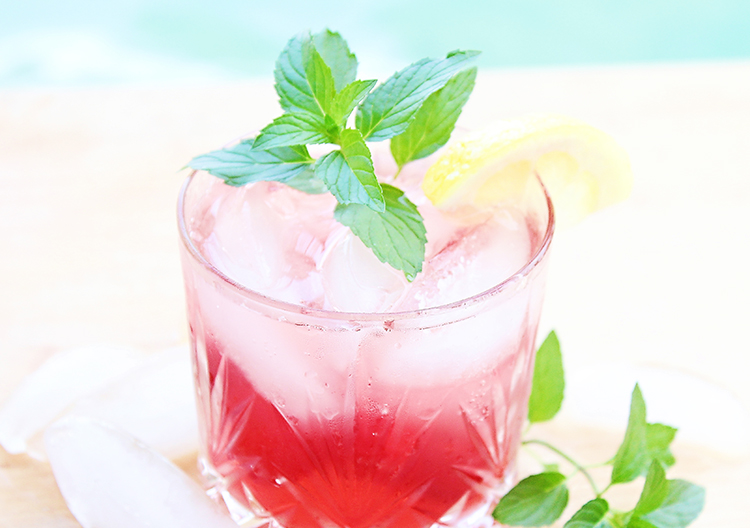 Cocktail in a rocks glass with cherry lemonade drink and a sprig of mint next to a pool for summer