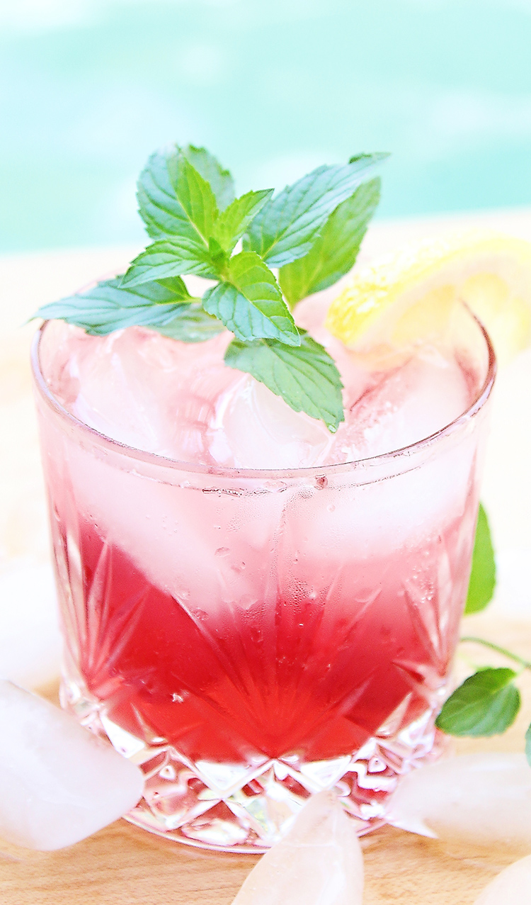 Cocktail in a rocks glass with cherry lemonade drink and a sprig of mint next to a pool for summer