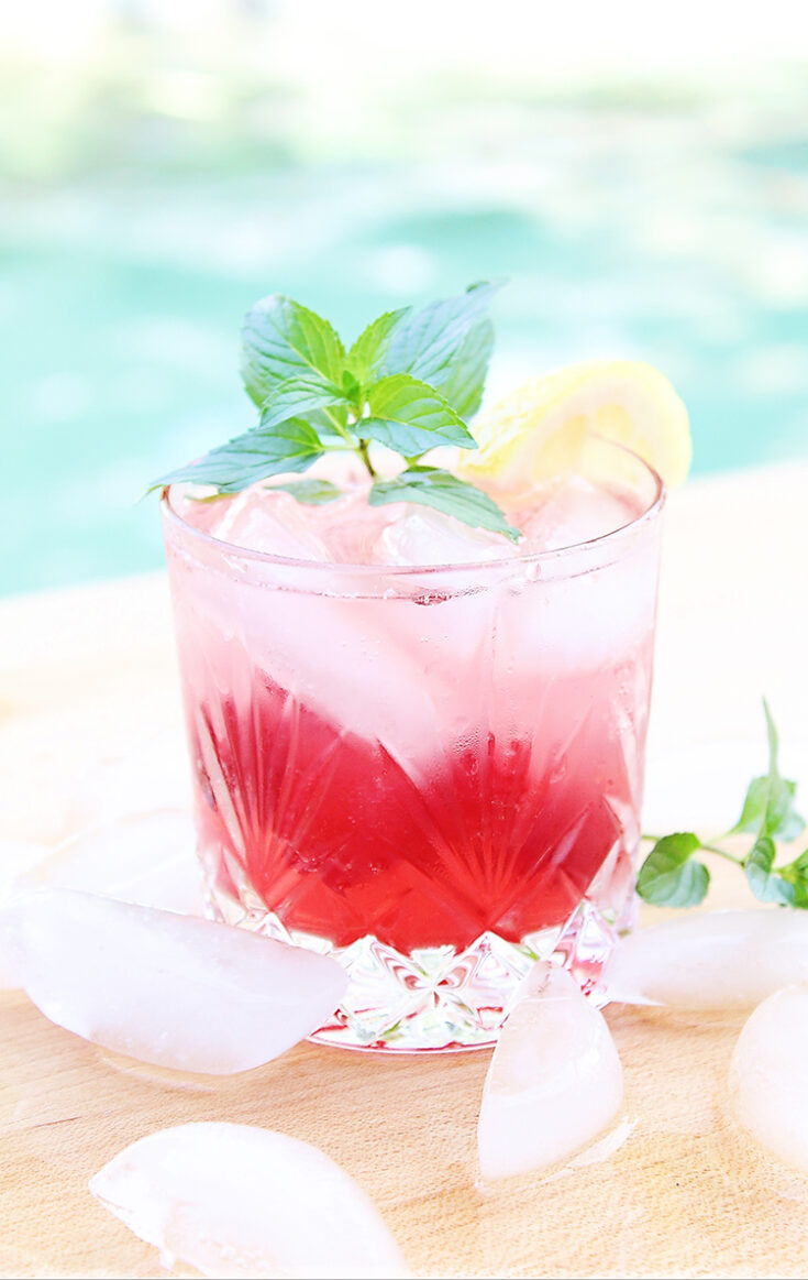 Cocktail in a rocks glass with cherry lemonade drink and a sprig of mint next to a pool for summer