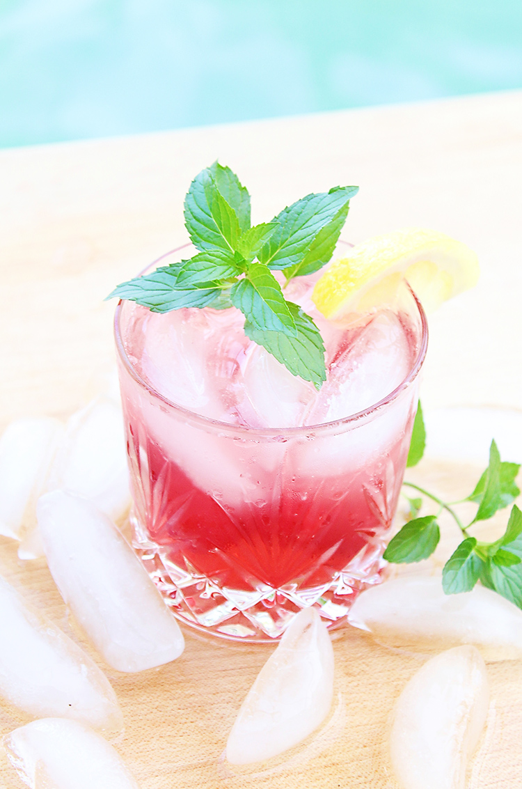 Cocktail in a rocks glass with cherry lemonade drink and a sprig of mint next to a pool for summer